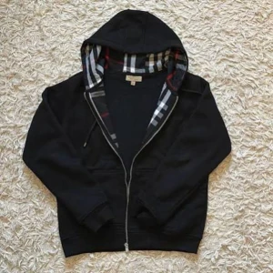 Svart hoodie från Burberry med dragkedja - Svart hoodie från Burberry med klassiskt rutigt foder i huvan. Tröjan har dragkedja framtill, snörning i huvan och ribbade muddar. Perfekt för dig som gillar stilrena och exklusiva plagg med en twist.