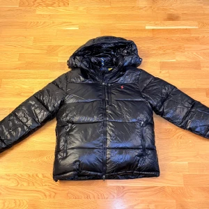 Svart pufferjacka från Polo Ralph Lauren - Säljer min svarta pufferjacka från Polo Ralph Lauren med stilren quiltad design och glansig finish. Jackan har huva, dragkedja framtill, flera praktiska fickor med dragkedja och den klassiska röda loggan broderad på bröstet – både varm och riktigt snygg i streetstil.  Storlek: L, men sitter även bra på dig som har M. Nypris: nästan 6000 kr (kvitto finns). Mitt pris: 2500 kr – säljs enbart för att jag vill köpa en annan jacka.