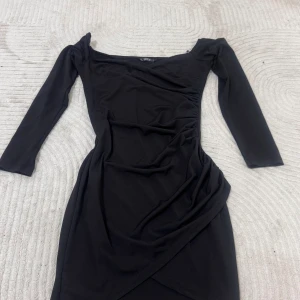 Svart omlottklänning från SHEIN - Svart figurnära klänning från SHEIN med omlottdesign och långa ärmar. Klänningen har en snygg offshoulder-ringning och lätt rynkade detaljer i sidan för en trendig look. Perfekt för dig som gillar stilrena och moderna plagg.
