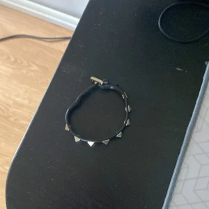 Valentino svart läderarmband med nitar - Svart armband i äkta läder från Valentino med silvriga pyramidformade nitar och spänne. Snyggt och edgy accessoar som ger en cool touch till din outfit. Justerbar längd med klassiskt Valentino-spänne. Snabb affär prioriteras. Mvh Anton