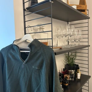 Grön stickad half zip från Bondelid - Mörkgrön stickad tröja från Bondelid med half zip och broderad logga på bröstet. Tröjan har ribbade muddar och hög krage, perfekt för kyliga dagar. Klassisk och stilren design som funkar till det mesta.