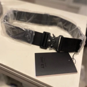 Svart bälte från DAY ET x SLS - Stilrent svart bälte från DAY ET x SLS med robust spänne i metall. Bältet har en bred, justerbar rem i syntetmaterial och en modern, minimalistisk look. Perfekt accessoar för att lyfta din outfit med en cool och urban känsla.