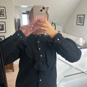 Svart skjorta med volang och knappar - Svart skjorta med volangdetalj längs bröstet och axlarna. Skjortan har klassisk krage, långa ärmar och knappar framtill. Materialet känns som bomull och modellen är normal i passformen. Jag säljer för att den blivit lite för liten för mig. Hör av dig om du vill ha fler bilder!
