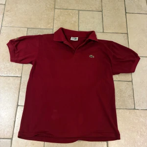 Vinröd Lacoste pikétröja XL - Vinröd pikétröja från Lacoste i storlek XL. Klassisk modell med krage, korta ärmar och broderad krokodil-logga på bröstet. Tillverkad i 100% polyester för en sportig känsla. Perfekt för dig som gillar stilren och tidlös design.