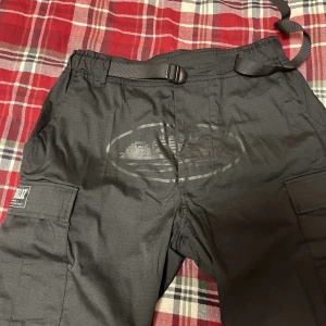 Svarta cargopants från Cortiez - Svarta cargopants från Cortiez med flera praktiska fickor på benen och patchdetaljer. Byxorna har justerbart bälte i midjan och är tillverkade i slitstark syntet. Perfekt för dig som gillar streetwear och funktionella detaljer.