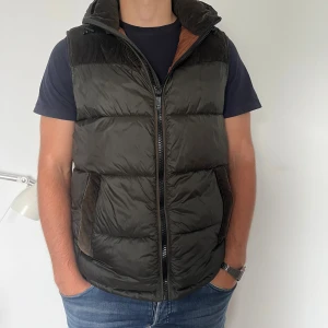 Mörkgrön dunväst med manchester  - Snygg mörkgrön dunväst med dragkedja och huva från Milestone. Varm och go väst som jag själv inte använder längre därav säljer jag den. Den är storlek L men passar M också