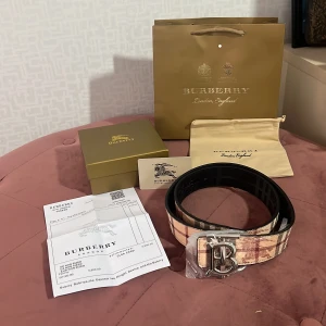 Burberry rutig skärp med silverdetalj - Exklusivt Burberry-bälte med klassiskt rutigt mönster i beige, brunt och rött. Skärpet har en bred design och en stor silvrig spänne med Burberrys ikoniska B-logga. Tillverkat i skinn och levereras med originalförpackning, dustbag och kvitto.