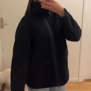 Svart teddyjacka med dragkedja - Mysig svart teddyjacka med hög krage och hel dragkedja framtill. Jackan har en loose passform och är perfekt för kyliga dagar. Den fluffiga ytan ger en trendig look och passar till de flesta outfits.