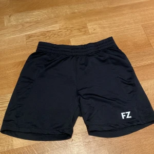  FZ Forza - Svarta träningsshorts - Badminton  - Svarta träningsshorts från FZ Forza med elastisk midja och snörning. Lätta och bekväma, perfekta för sport eller gym. Diskret FZ-logga på ena benet. Tillverkade i ett syntetmaterial som andas.