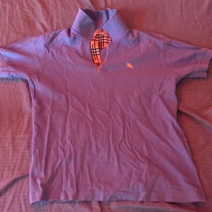 Blå Burberry pikétröja med rutig krage - Säljer en blå pikétröja från Burberry med klassisk rutig detalj vid kragen och gul broderad logga på bröstet. Tröjan har korta ärmar och är gjord i mjuk bomull. Perfekt för dig som vill ha en stilren och ikonisk look.