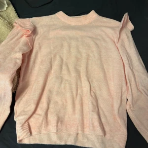 Ljusrosa stickad tröja med volang - Superfin ljusrosa stickad tröja med volangdetaljer på axlarna. Tröjan har rund halsringning, långa ärmar och ribbade muddar vid ärmslut och nederkant. Perfekt för dig som vill ha en söt och trendig look.