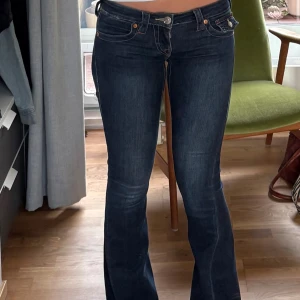 True Religion Joey Low Rise Flare Jeans - Säljer ett par mörkblå True Religion Joey jeans med låg midja och flare-ben. Snygga kontrastsömmar och ikoniska fickdetaljer med lock och knapp bak. Klassisk femficksmodell i stretchig denim som sitter tajt upptill och släpper ut vid vaden. Sömmen vid nedre delen av byxan har gått av som man kan se på bild 5 men absolut inget man tänker på.