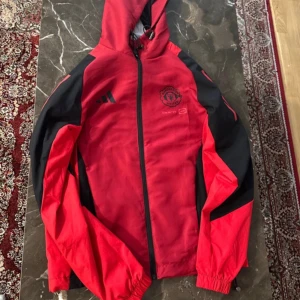 Manchester United vindjacka Adidas röd - Säljer en röd och svart Manchester United vindjacka från Adidas med huva och dragkedja. Jackan har elastiska muddar, snörning i huvan och klassiska Adidas-ränder på ärmarna. Perfekt för dig som vill representera ditt lag med stil.