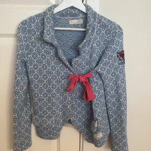 Blåvit kofta Odd Molly med rosett - Superfin blåvit mönstrad kofta från Odd Molly med lång ärm, broderad ros på ärmen och texten 'Rise Above'. Koftan har en rosa rosett framtill och är stickad i mjukt material. Perfekt för dig som gillar unika detaljer och snygg passform.