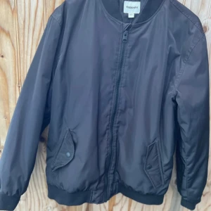 Svart bomberjacka från Kappahl - Svart bomberjacka från Kappahl i storlek 152. Jackan har klassisk bombermodell med ribbade muddar, dragkedja framtill och två fickor med knappstängning. Perfekt för dig som gillar en stilren och enkel look.