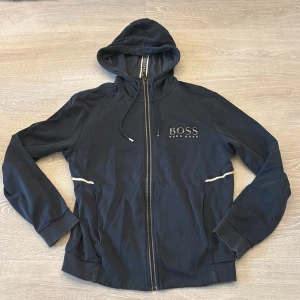 Marinblå hoodie från Hugo Boss - Snygg marinblå hoodie från Hugo Boss med dragkedja och huva. Regular fit med broderad logga på bröstet och vita detaljer längs sidorna. Mjukt material och ribbade muddar vid ärmslut och nederkant. Perfekt för chill dagar.