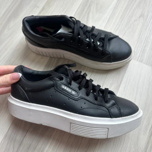 Svarta Adidas sneakers - Snygga svarta Adidas sneakers med vit platåsula och klassisk snörning. Skorna har en rund tå och är tillverkade i skinn med diskret perforering på sidorna. Adidas-logga i vitt på sidan ger en clean och modern vibe. Perfekta för dig som gillar streetstyle och vill ha extra höjd.