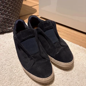 Arigato skor - Svarta slip-on sneakers med ovandel i mjuk mocka och bred resår över vristen. Skorna har en vit platt sula och rund tå, vilket ger en clean och stilren look. Perfekta för dig som gillar minimalistisk stil och vill ha något enkelt att dra på sig. Storlek 44