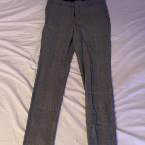 Grå rutiga kostymbyxor herr - Snygga grå kostymbyxor med diskret rutigt mönster. Klassisk rak passform med fickor fram och bak, samt hällor för bälte. Perfekta för en stilren look och passar till skjorta eller t-shirt.