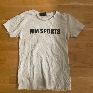 Vit t-shirt från MM Sports - Vit t-shirt från MM Sports med svart tryck på bröstet. Klassisk rund halsringning och korta ärmar. Tillverkad i mjuk bomull, perfekt för en sportig och avslappnad stil.