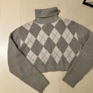 Grå argyle polotröja Pull&Bear - Supermysig grå polotröja från Pull&Bear med klassiskt argylemönster i vitt, grått och ljuslila. Tröjan är stickad, har lång ärm och croppad passform. Perfekt för lager på lager och snygg till både jeans och kjol.