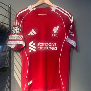 Liverpool röd fotbollströja Adidas - Säljer en röd Liverpool fotbollströja från Adidas med vita detaljer och tryck. Tröjan har korta ärmar, klubbmärke och sponsortryck på bröstet samt Champions League-märke på ärmen. Namn och nummer 'Ekitike 22' på ryggen. Materialet är lätt och ventilerande polyester. Helt ny med tagg en replika 