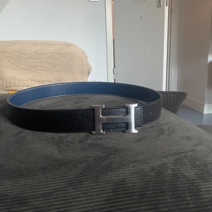 H svart skärp med H-spänne - Säljer ett stilrent svart skärp från H med klassiskt H-spänne i silver. Bältet är gjort i skinn och har en blå insida som ger en snygg kontrast. Perfekt accessoar för att lyfta din outfit.