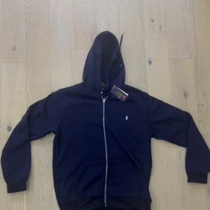 Mörkblå Ralph lauren zip - Mörkblå hoodie från Polo Ralph Lauren med dragkedja framtill, klassisk huva och fickor. Liten vit logga broderad på bröstet. Perfekt för en chill och stilren look.