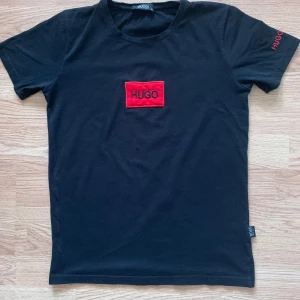 Svart t-shirt Hugo Boss röd logga - Svart t-shirt från Hugo Boss med röd broderad HUGO-logga på bröstet och HUGO BOSS-broderi på ärmen. Klassisk passform och rund hals. Tillverkad i mjuk bomull för en skön känsla.