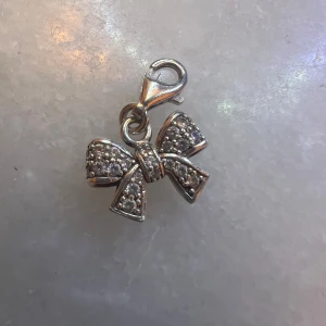 Thomas Sabo silver rosettberlock - Superfin berlock från Thomas Sabo i form av en rosett, tillverkad i silver och dekorerad med gnistrande stenar. Perfekt att hänga på armband eller halsband för en söt och glittrig detalj. Lätt att fästa med karbinhake.