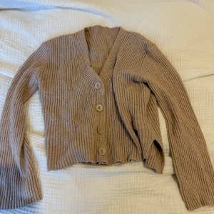 Beige ribbad kofta från SHEIN - Säljer en beige ribbad kofta från SHEIN med v-ringning och fyra stora knappar framtill. Koftan har långärmad passform och är croppad i modellen, vilket ger en trendig look. Perfekt att slänga över en t-shirt eller linne för extra stil.