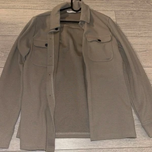 Beige overshirt från Only & Sons - Snygg beige overshirt från Only & Sons med klassisk krage och två stora bröstfickor med knappar. Långärmad modell i mjukt material, perfekt att slänga över en t-shirt. Enkel och stilren design som funkar till många olika looks.