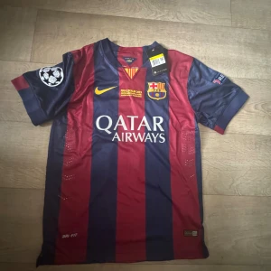 FC Barcelona Neymar Jr 11 Finaltröja - Säljer en officiell FC Barcelona matchtröja från Champions League-finalen 2015 med Neymar Jr och nummer 11 på ryggen. Tröjan är randig i mörkblått och rött, har korta ärmar, broderat klubbmärke och Nike-logga, samt tryckta sponsorer. Materialet är lätt polyester med Dri-FIT-teknologi.