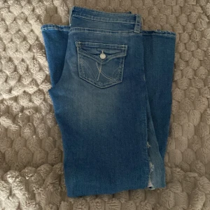 Blå bootcut jeans med slitningar - Säljer ett par blå bootcut jeans med slitningar på benen och snygga detaljerade bakfickor med knappar. Jeansen har klassisk femficksdesign, markerade sömmar, lågmidjade och är tillverkade i denim. Perfekta för dig som gillar en retro vibe och vill sticka ut lite extra.