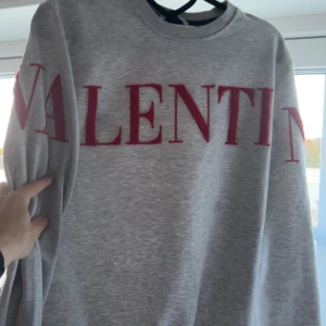 Grå sweatshirt från Valentino - Snygg grå sweatshirt från Valentino med stort rött logotryck över bröstet och ärmarna. Tröjan har rund halsringning och långa ärmar. Perfekt för dig som vill ha en stilren men ändå statement look. LITEN FLÄCK FINNS PÅ TRÖJAN, skriv privat för tydligar bilder