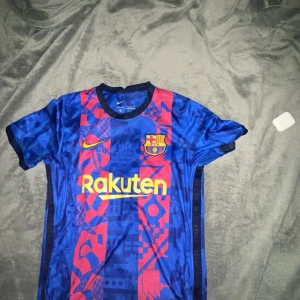 FC Barcelona Nike Dri-FIT matchtröja M - Säljer en FC Barcelona matchtröja från Nike i Dri-FIT-material. Tröjan är blå med röda grafiska mönster och gula detaljer, klubbmärke på bröstet och Rakuten-tryck framtill. Korta ärmar och sportig passform, perfekt för fotbollsälskare.