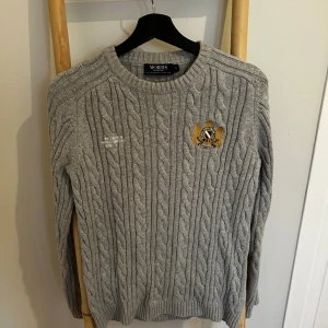 Grå kabelstickad tröja från Morris - Kabelstickad grå tröja från Morris med broderad patch på bröstet i gult och svart. Tröjan har rund halsringning, ribbade muddar och är långärmad. Perfekt för dig som gillar klassisk och stilren look.