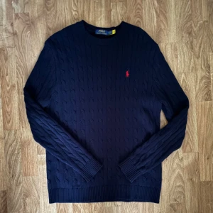 Mörkblå stickad tröja från Polo Ralph Lauren - Kabelstickad mörkblå tröja från Polo Ralph Lauren med klassisk röd broderad logga på bröstet. Tröjan har rund halsringning, ribbade muddar och är långärmad. Perfekt för lager-på-lager under kyliga dagar.