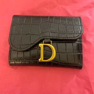 Svart plånbok med gulddetalj  - Snygg svart plånbok med krokopräglad yta och stor guldfärgad D-detalj framtill. Insidan har plats för kort och sedlar, med en cool kontrast i gult. Perfekt accessoar för dig som gillar lyxiga detaljer och stilren design.