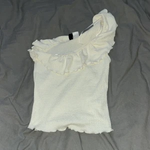 Krämvit oneshoulder topp från H&M -  krämvit oneshoulder topp från H&M i storlek XS. Toppen har en volang längs halsringningen och är tillverkad i ett mjukt, lätt strukturerat material💕