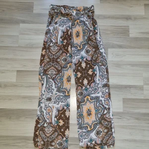 Mönstrade vida byxor från Amisu - Säljer ett par supercoola vida byxor från Amisu i storlek 40. Byxorna har ett färgstarkt paisleymönster i brunt, gult, turkost och vitt samt knytband i midjan. De är tillverkade i ett lätt och luftigt material, perfekta för en chill och trendig look.
