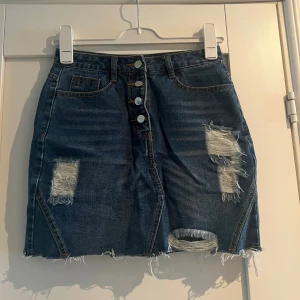 Mörkblå jeanskjol med slitningar från Shein S - Trendig mörkblå jeanskjol från Shein i kort modell med rå kant nertill och slitna detaljer framtill och baktill. Kjolen har knappar framtill, klassiska fickor storlek S, aldrig använd 
