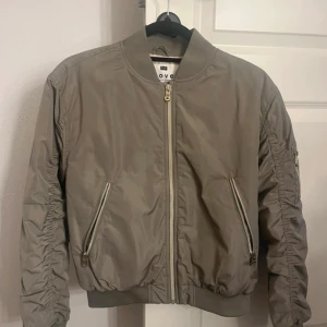 Beige bomberjacka F&F - Snygg beige bomberjacka från F&F  med dragkedja framtill och två snedställda fickor med dragkedja. Jackan har ribbade muddar vid ärmslut och midja samt detaljerad rynkning längs ärmarna. Perfekt för dig som vill ha en stilren och trendig look.