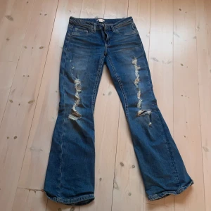 Blå bootcut jeans från Gina Tricot - Snygga blå jeans från Gina Tricot med bootcut passform och slitna detaljer framtill. Klassiska femficksmodellen med dekorativa sömmar på bakfickorna och knappstängning. Perfekt för dig som gillar en avslappnad och trendig stil.