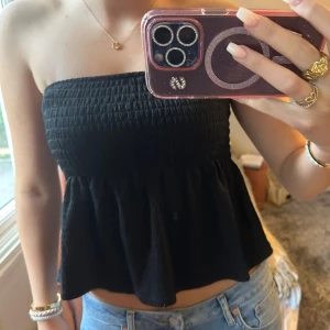 Svart bandeau tubtopp med volang - Säljer en svart bandeau tubtopp med smockad överdel och volangig nederdel. Toppen är axelbandslös och har en luftig, flowy passform som är perfekt till jeans eller kjol. Materialet känns mjukt och lätt, och toppen är superenkel att styla för en trendig look.