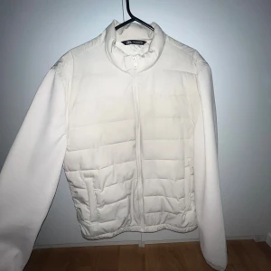 Vit pufferjacka från Zara - Säljer en stilren vit pufferjacka från Zara med quiltad framsida och dragkedja. Jackan har hög krage, två fickor och långärmade släta ärmar. Perfekt för dig som vill ha en clean look under kyliga dagar.