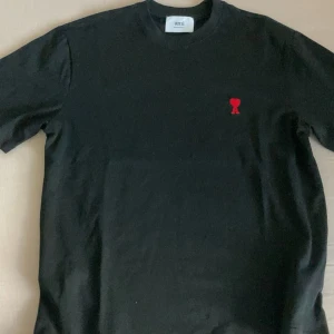 Svart t-shirt från Ami med rött hjärta - Svart t-shirt från Ami Alexandre Mattiussi med klassisk passform. T-shirten har ett broderat rött hjärta med bokstaven A på bröstet. Tillverkad i mjuk bomull och har rund halsringning samt korta ärmar. Perfekt för en clean och stilren look.