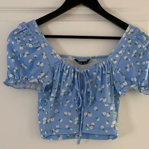 Blommig blå croppad blus från SHEIN - Supersöt ljusblå blus från SHEIN med små vita blommor och gröna detaljer. Croppad modell med puffiga korta ärmar, rynkade kanter och knyt framtill. Perfekt för dig som gillar romantisk och trendig stil.