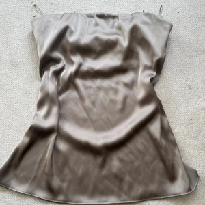 Champagnefärgad axelbandslös topp - Säljer en stilren axelbandslös topp/klänning från Zara i champagnefärgad satin. Den har en rak och loose passform och är tillverkad i ett glansigt, mjukt material som ger en lyxig känsla. Aldrig använd