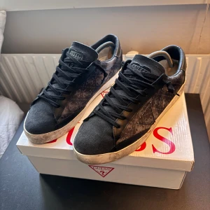 Svarta sneakers från Guess, storlek 42 - Snygga svarta sneakers från Guess med diskret mönstrad sida och logga på plösen. Skorna har svarta snören, vit sula och ovandel i en mix av mocka och syntet. Perfekta för dig som gillar stilrena men ändå unika detaljer.